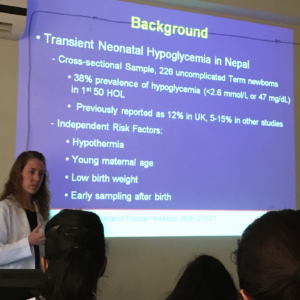 Person presenting a slide presentation in a classroom or conference setting, with audience members visible in the foreground.